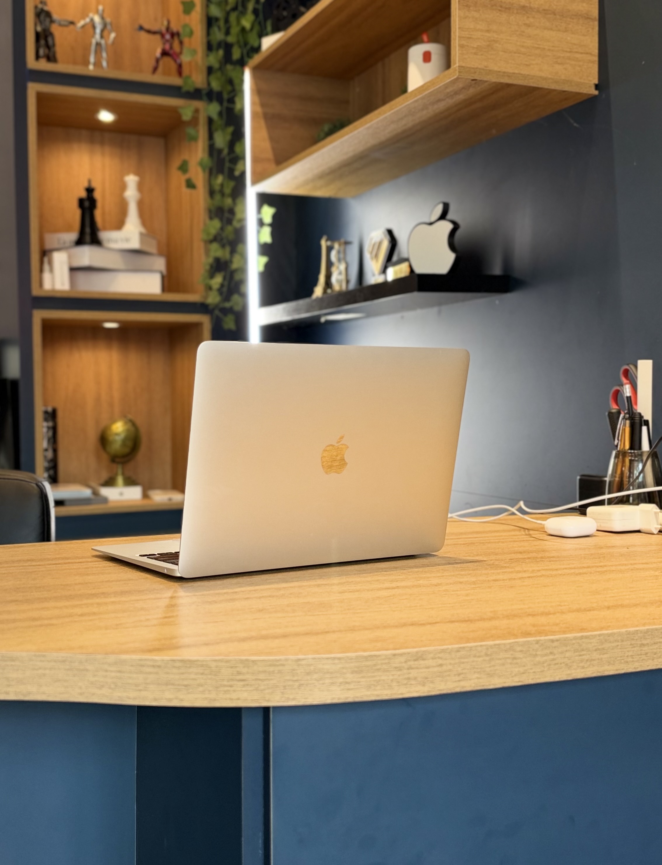 MacBook na bancada de madeira da MJ Store, com parede azul e prateleiras iluminadas